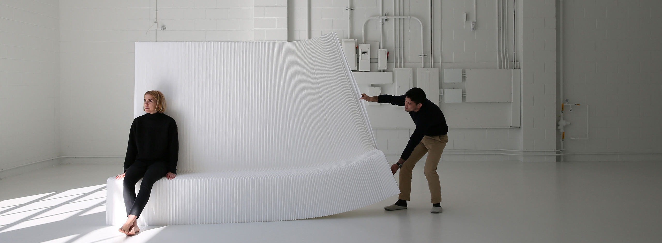 a man tries to stretch open a white textile benchwall while a woman sits on it and weighs it down