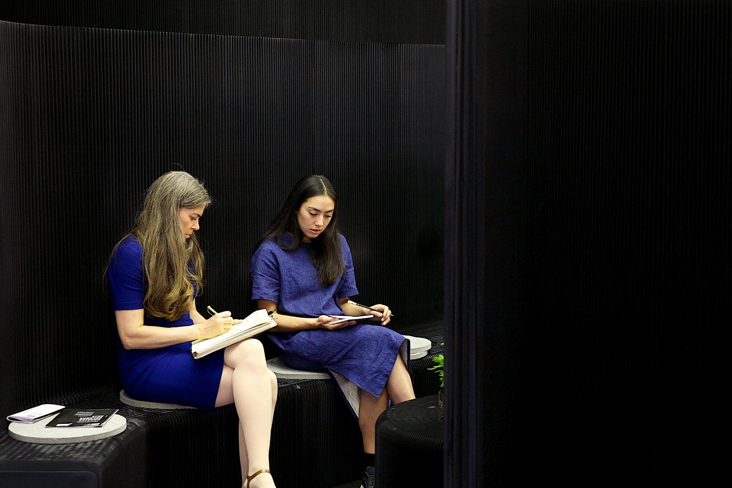 Two women in blue dresses sit on wool felt pads atop a black benchwall